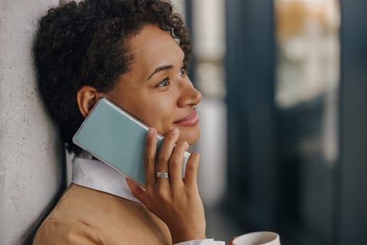 Smiling female manager with coffee cup is talking phone while standing on modern office background