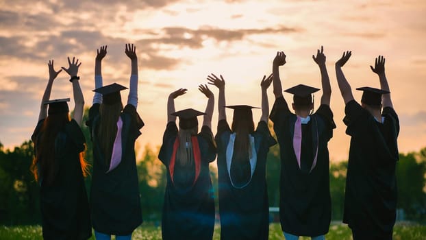 Silhouettes of graduates in black robes waving their arms against the evening sunset