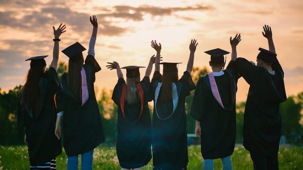 Silhouettes of graduates in black robes waving their arms against the evening sunset