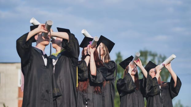 Cheerful graduates on a sunny day look through diplomas like a telescope