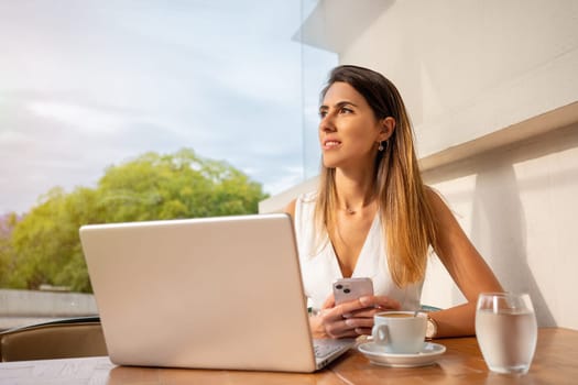 Businesswoman sitting in an outdoor cafe using laptop. Hispanic adult woman sitting in an online meeting in street cafe, drinking coffee. Female freelancer in white dress working or studying on laptop