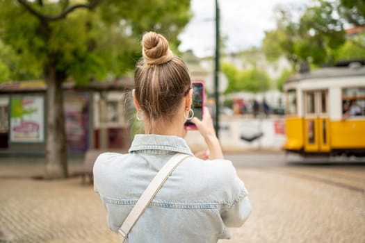 Happy traveler girl in Lisbon old city. Tourist woman taking picture tramway in Portugal. Holiday vacation in european city. Female tourist making photo using smartphone back view