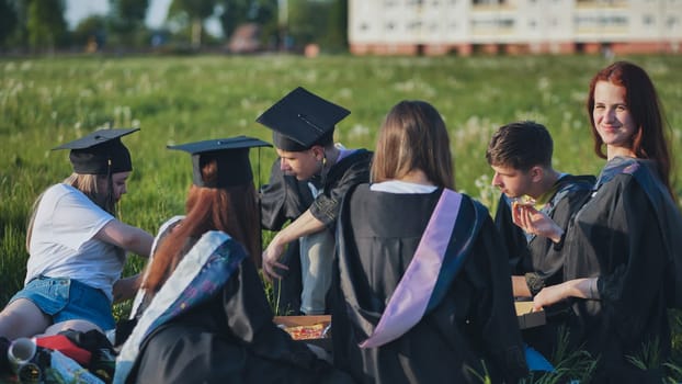 Graduates in black suits eating pizza in a city meadow