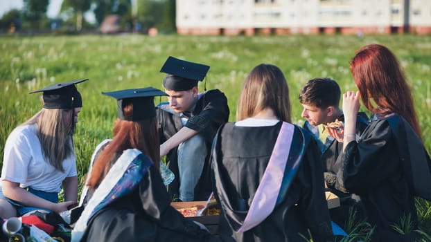 Graduates in black suits eating pizza in a city meadow