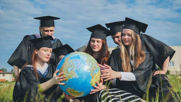 Graduates in black robes examine a geographical globe sitting on the grass