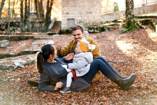 Smiling dad sits near mom with a little girl on her belly on fallen leaves in the forest. High quality photo