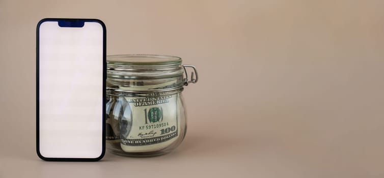 Vertical White screen on modern mobile phone in background of glass jar full of American currency dollar banknotes on beige background. Cope space for text. Advertisement for application website. Concept of money economy banks and finances