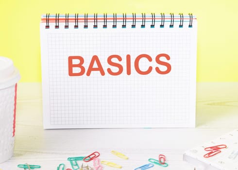 BASICS the word is written on a blank sheet in a notebook standing on a table on a yellow background