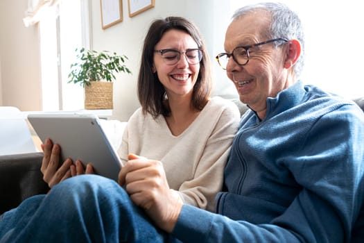Happy senior man and his young daughter laughing together using digital tablet sitting on sofa at home. Family members sharing technology.