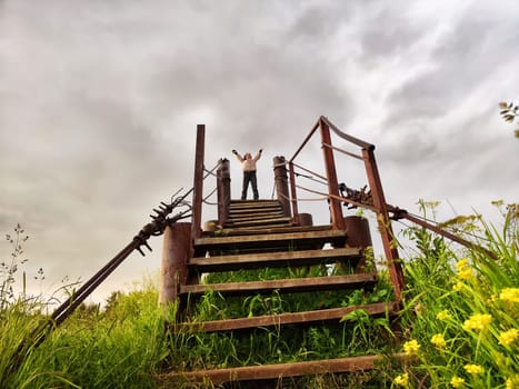 High, old, rusty iron staircase and tiny statue of girl or woman at the top against backdrop of gray, gloomy, thunderous clouds. Concept of an unusual, fantastic journey, surrealism, strange world