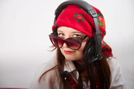 Portrait of young girl in red scarf, large headphones with microphone and black glasses. Woman who is radio or television presenter in workplace. Funny female telecom operator. Freelancer at work