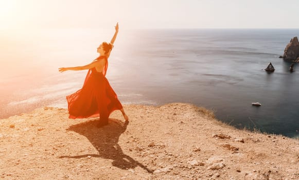 Side view a Young beautiful sensual woman in a red long dress posing on a rock high above the sea during sunrise. Girl on the nature on blue sky background. Fashion photo.