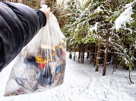 Bag of garbage in hand of woman and snow in forest on the background. A girl cleans up the trash after picnic. A tourist is going to throw garbage in nature. The concept of environmental pollution