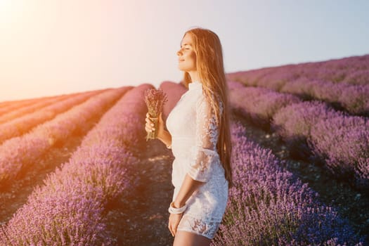 Close up portrait of young beautiful woman in a white dress and a hat is walking in the lavender field and smelling lavender bouquet.