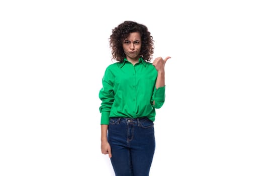 young caucasian brunette woman with curled hair dressed in a green shirt points her finger towards copy space. advertising concept.