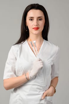 the girl cosmetologist holds two brushes in her hands on a white background, the girl is a beautician in white disposable gloves.