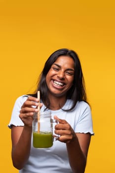 Vertical portrait of happy young beautiful Indian woman with green smoothie on yellow background looking at camera. Healthy lifestyle concept.