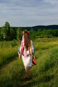 The girl, with her back turned and dressed in Ukrainian national clothes, walks barefoot in the field, wearing a wreath with ribbons on her head.