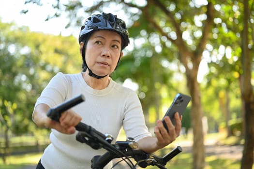 Senior woman in sports clothing riding bicycle at the park and using mobil phone