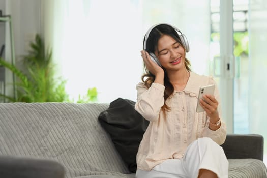 Cheerful young woman listening to music in headphones and using smartphone on couch