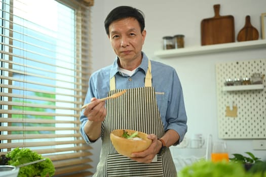 Happy Asian senior man eating healthy vegetable salad in kitchen. Healthy lifestyle, dieting and nutrition concept
