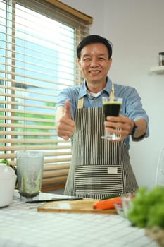 Cheerful middle age man holding glass of green smoothie and showing thumbs up