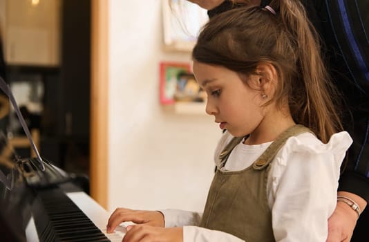 Adorable Caucasian little child girl playing piano under the guidance of her teacher, holding a paper with musical notes and learning new music composition or classic melody. People, Arts and culture