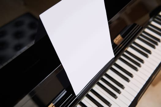 Close-up of old black wooden piano with ivory and ebony piano keys and white paper sheet with copy space for notes or advertising text. View from above