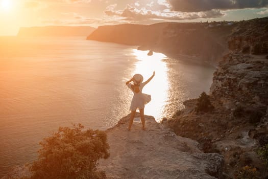 woman sea travel. photo happy woman with hat in white dress standing on the shore on a hill overlooking the sea. golden hour, silhouette of a woman at sunset on the mountain