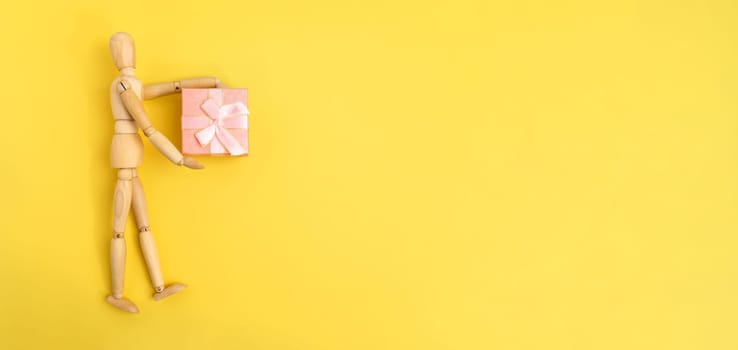 Wooden figurine holding a pink gift box. Yellow background, copy space. Banner