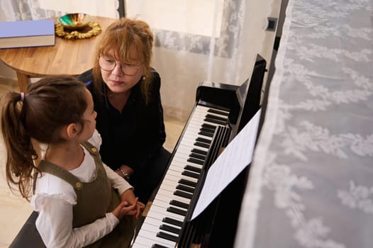 Overhead view of inspired Caucasian beautiful little child girl, focused engaged on learning playing grand piano with female teacher musician and pianist during individual music lesson at home