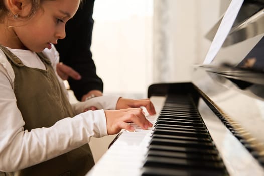 Caucasian little child girl playing grand piano, touching piano keys, performing musical rhythm during individual music lesson with her teacher, a professional female musician pianist at home