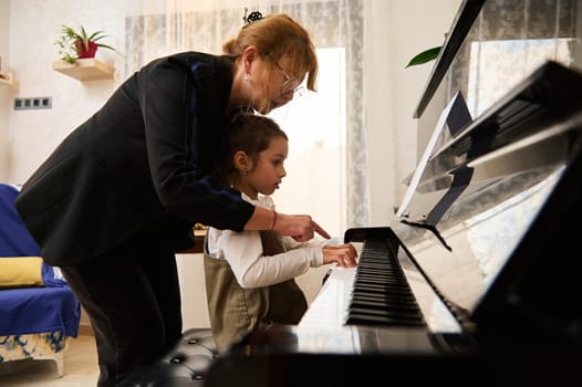 Mature woman, pianist teacher giving piano lesson to a little school girl, teaching and showing the position of fingers on piano keys, playing music together on old piano at cozy home interior