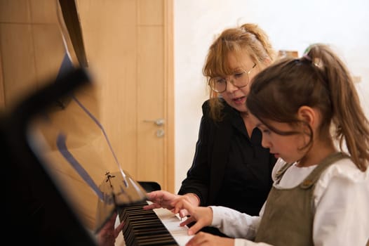 Caucasian adorable elementary age smart school girl having piano lesson with mature female teacher, pianist. Little child girl playing grand piano.