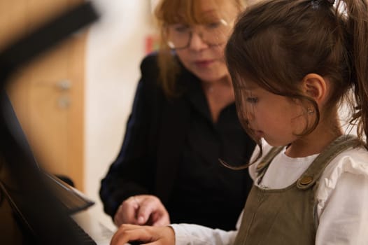 Caucasian adorable smart school girl having piano lesson with mature female teacher, pianist. Little child girl playing grand piano.