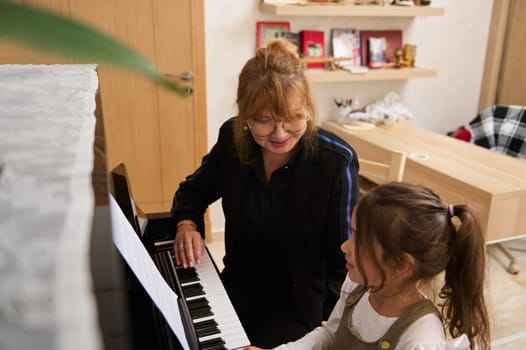 Beautiful mature woman, pianist teacher giving music lesson to a smart child girl, teaching her playing on grand piano during individual class at home. People, Hobbies and leisure. Music and arts