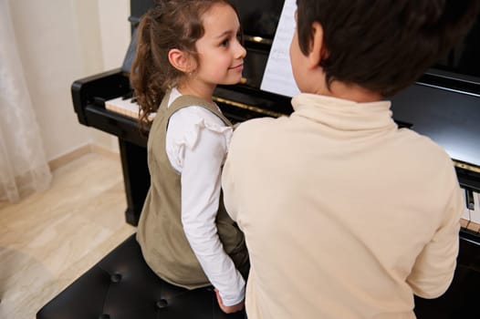 Rear view of two kids playing grand piano together, creating rhythm of classical melody during individual music lesson indoor.