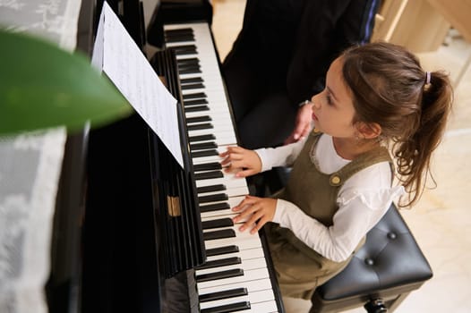 Top view of a little pianist girl playing piano, composing a melody under the guidance of her teacher during individual music lesson at home. Children. Education. Lifestyles. Hobbies and entertainment