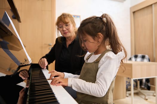 Kid girl playing piano, creating music, touching piano keys with her fingers, feeling the rhythm of music, practicing music on chord instrument with her teacher during individual lesson