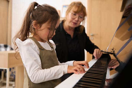Mature female musician, a music teacher, pianist, teaching piano, explaining to a little kid girl student the correct position of fingers on the piano keys. People. Arts, culture and entertainment