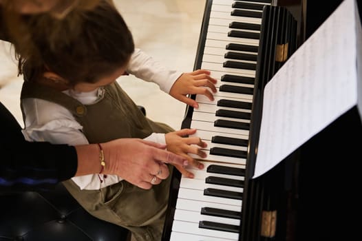 Top view music teacher, pianist holding the hands of a little child girl, teaching piano lesson to a student, showing her the correct finger position. Kids education, artistic skills development