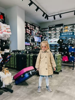 Little girl in sunglasses stands in a store holding a travel suitcase by the handle. High quality photo