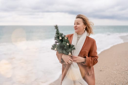 Blond woman Christmas sea. Christmas portrait of a happy woman walking along the beach and holding a Christmas tree in her hands. She is wearing a brown coat and a white suit