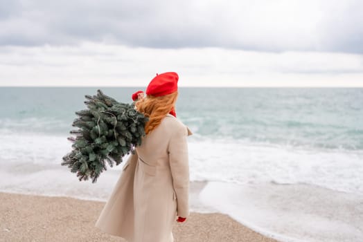 Redhead woman Christmas tree sea. Christmas portrait of a happy redhead woman walking along the beach and holding a Christmas tree on her shoulder. She is dressed in a light coat and a red beret