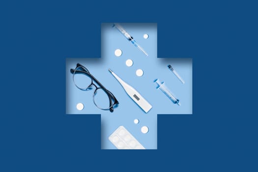 Medical classic blue background with different accessories: syringe, tablets, thermometer, glasses lying in a carved medical cross.