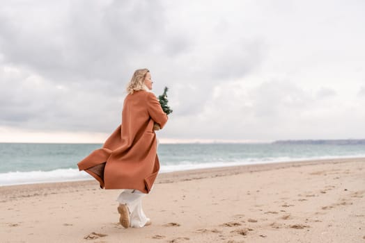 Blond woman Christmas sea. Christmas portrait of a happy woman walking along the beach and holding a Christmas tree on her shoulder. She is wearing a brown coat and a white suit