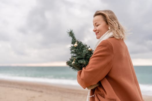 Blond woman Christmas sea. Christmas portrait of a happy woman walking along the beach and holding a Christmas tree on her shoulder. She is wearing a brown coat and a white suit