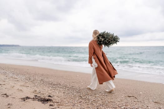 Blond woman Christmas sea. Christmas portrait of a happy woman walking along the beach and holding a Christmas tree on her shoulder. She is wearing a brown coat and a white suit