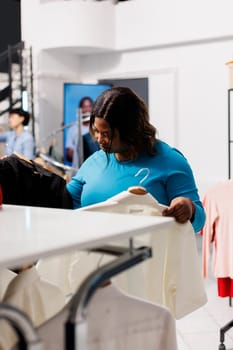 African american woman looking at hangers with elegant shirt, checking items fabric in modern boutique. Shopaholic clients buying new fashion collection, shopping for casual wear in clothing store