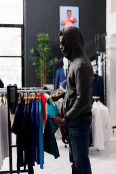 African american man looking aside while robbing clothing store, checking racks with trendy clothes. Thief trying to steal fashionable merchandise, wearing sunglasses and hood not to be recognized